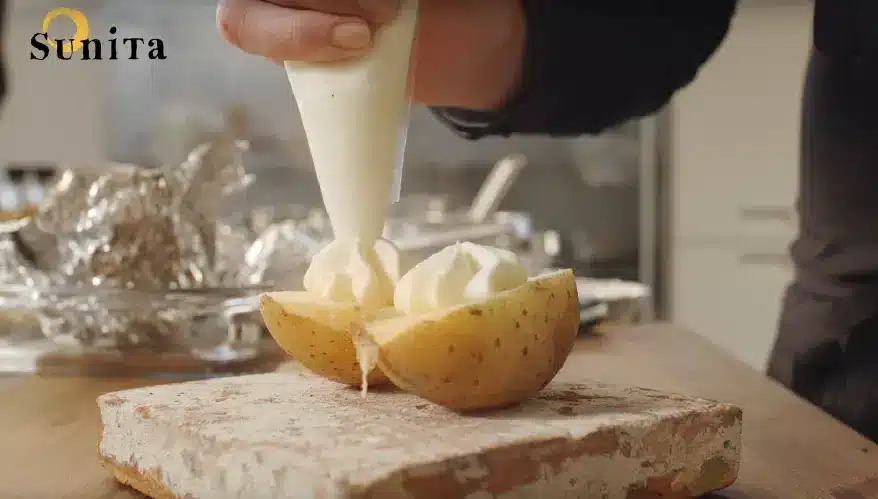 Opengesnede gekookte aardappel op een plateau waarop een witte crème uit spuitzak wordt gespoten
