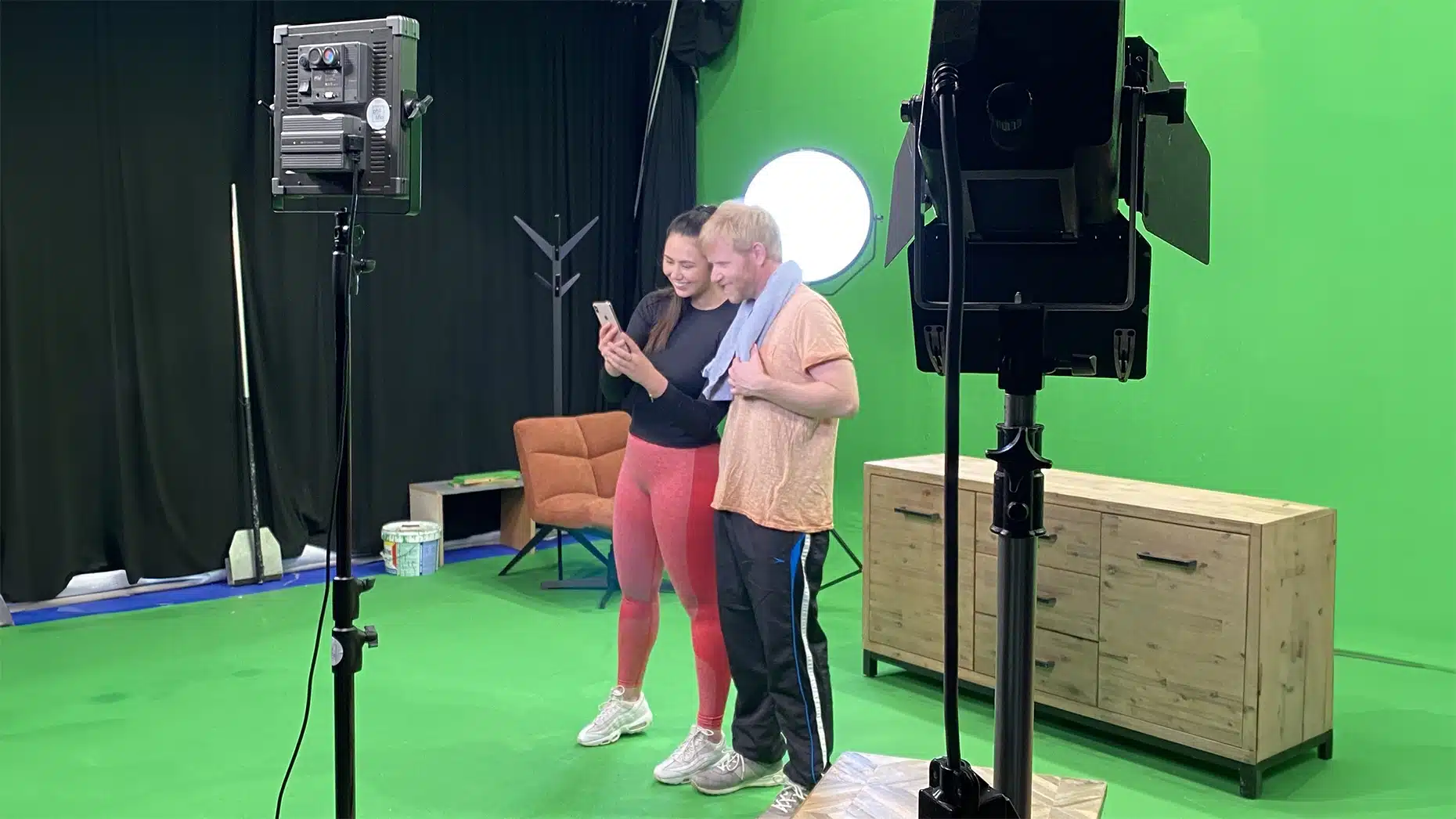 Een meisje en jongen in sportkleding staan in een green screen studio naast elkaar en lachen naar de mobiele telefoon die het meisje vast heeft
