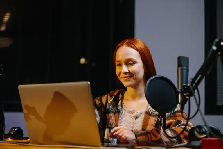Een vrouw met rood haar glimlacht naar het computerscherm waar een studio microfoon naast staat